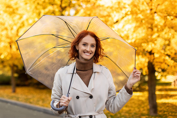 Happy caucasian millennial woman with red hair in raincoat with umbrella enjoys rain, weather and yellow leaves outdoor, free space. Walk in city park at spare time, human emotions and fun alone - Zdjęcie, obraz
