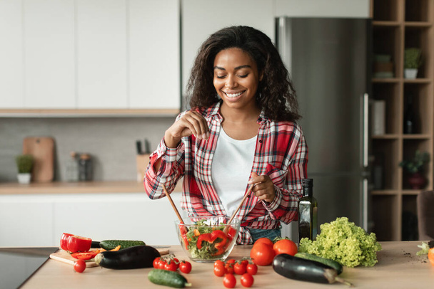 Domácí jídlo a zdravotní péče. Veselá černá mladá žena kuchařka v neformální soli salát z ekologické zeleniny v moderním kuchyňském interiéru. Vaření vegetariánské jíst doma, správné výživy a životního stylu - Fotografie, Obrázek