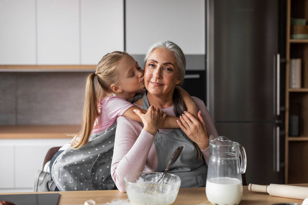 Örülök, hogy boldog kaukázusi kislány ölel és csókol az idős nagymama arcába, tésztát készít a modern konyha belsejében való sütéshez. Édességek főzés lecke együtt otthon, szerelem és kapcsolat - Fotó, kép