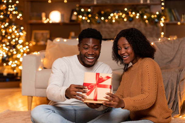 Σοκαρισμένο ευτυχισμένο νεαρό μαύρο ζευγάρι ανοίγει κουτί με δώρο στο άνετο σαλόνι εσωτερικό με χριστουγεννιάτικο δέντρο με φώτα και εορταστική διακόσμηση. Έκπληξη δώρο για το ζευγάρι στο γιορτάσουν το νέο έτος μαζί στο σπίτι - Φωτογραφία, εικόνα