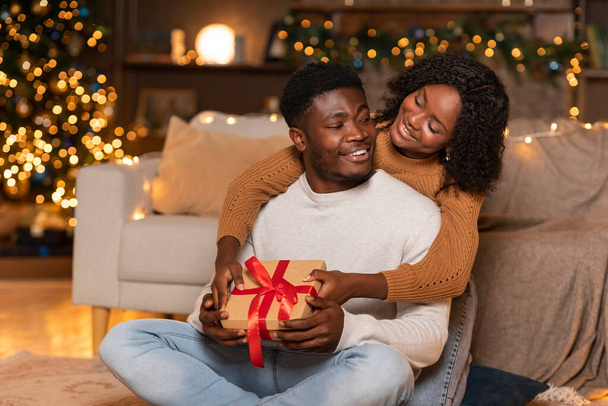 Wesoły młody Afrykańska kobieta przytulić faceta z pudełka prezentów w przytulnym salonie wnętrze z choinki z lampkami i wystrojem. Niespodzianka i obecny na zimowych wakacjach, cieszyć się obchodami Nowego Roku - Zdjęcie, obraz