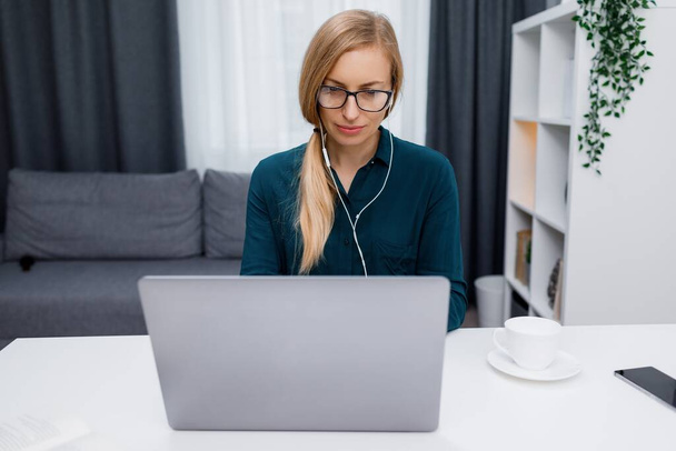 Εστιασμένη ώριμη γυναίκα σε ακουστικά πληκτρολογώντας σε ασύρματο φορητό υπολογιστή, ενώ κάθεται στο γραφείο στο σπίτι. Τηλεργασία και τεχνολογική έννοια. - Φωτογραφία, εικόνα