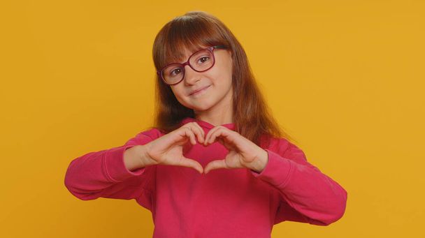 I love you. Smiling young preteen child girl kid makes heart gesture demonstrates love sign expresses good feelings and sympathy. Little toddler children isolated alone on studio yellow background - Fotografie, Obrázek