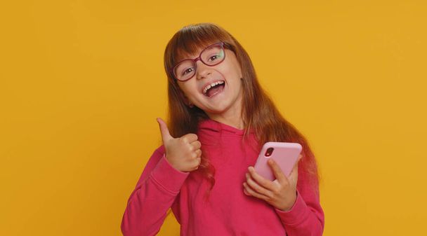 Young preteen child girl kid use mobile smartphone typing browsing say Wow yes found out great big win good news celebrate doing winner gesture. Little toddler children on studio yellow background - Foto, immagini