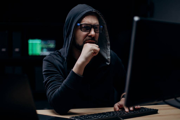 Attentive computer hacker on black hoodie breaking into corporate data while sitting at secret room at night. Mature bearded man in eyeglasses typing on keyboard of modern pc. - Foto, Imagen