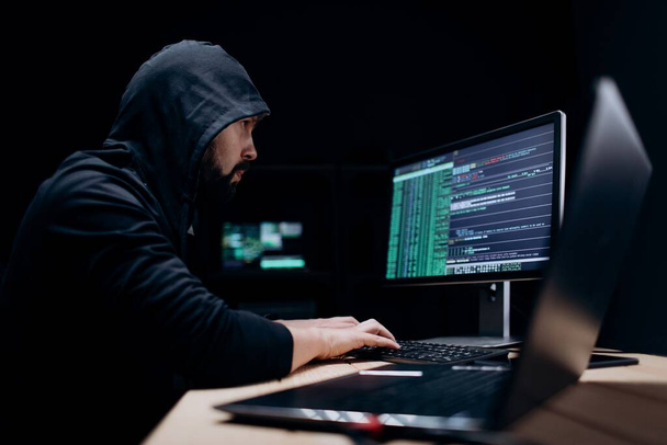 Side view of mature bearded man sitting at dark room and coding on modern pc and laptop. Caucasian male IT specialist destroying information on government servers. - Photo, Image