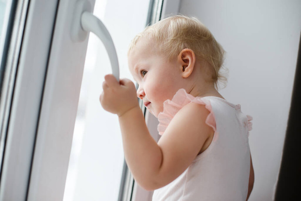 baby is trying to open the window in the apartment. No lock on the window. Dangerous home for a baby. Fall safety for children. The child stands on the windowsill of a closed window and tries to open the window sash without a lock - Foto, imagen