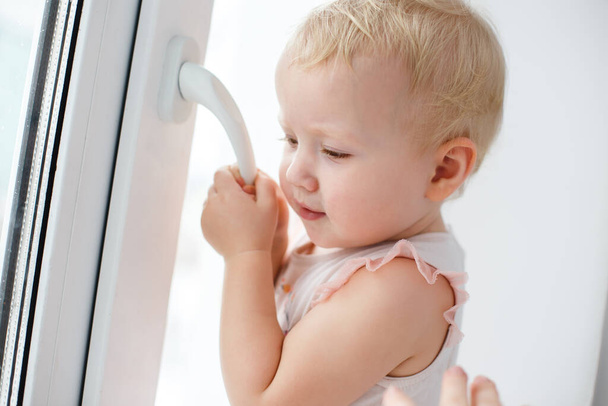 Προστατεύει τα παιδιά από το να πέσουν από τα παράθυρα. Ένα δραστήριο κοριτσάκι ανοίγει προσεκτικά ένα μεταλλικό-πλαστικό παράθυρο στο σπίτι.. - Φωτογραφία, εικόνα