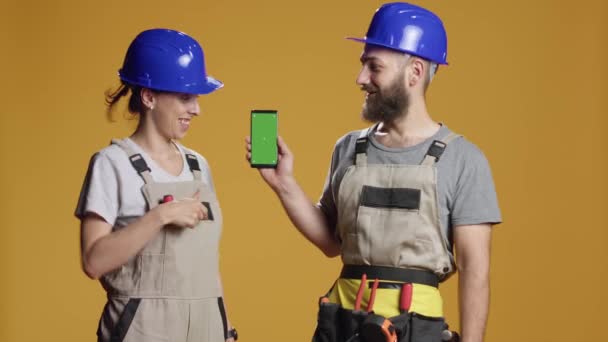 Portrét stavebních dodavatelů ukazujících na smartphone se zelenou obrazovkou ve studiu, pracujících na výstavbě. Držení izolované šablony mockup s prázdným kopírovacím prostorem chroma kláves. - Záběry, video