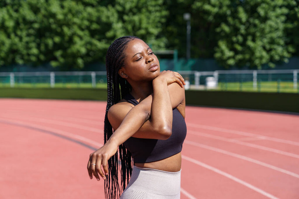 African American sportswoman with long braids enjoys training in sunny morning. Young woman stretches hands and shoulders standing on red track of stadium against lush green trees in city park - Fotografie, Obrázek