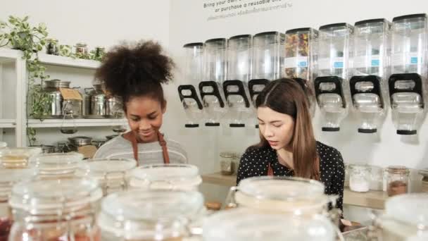 Twee jonge vrouwelijke winkeliers werken in een navulwinkel en controleren de voorraad biologische producten in herbruikbare potten, nul afval en milieuvriendelijke kruidenierswinkel. Thaise tekst op de muur betekent breng uw eigen containers - Video