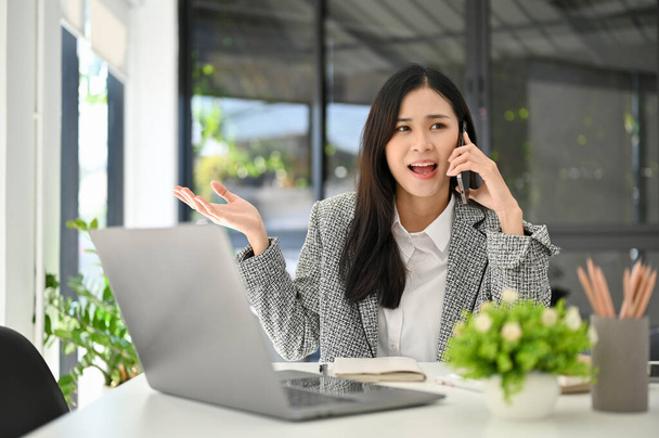 Όμορφη νεαρή Ασιάτισσα επιχειρηματίας ή γυναίκα στέλεχος λογαριασμού μιλώντας στο τηλέφωνο με τον πελάτη της, που ασχολείται με επιχειρήσεις μέσω της κλήσης, ενώ εργάζεται στο γραφείο της. - Φωτογραφία, εικόνα