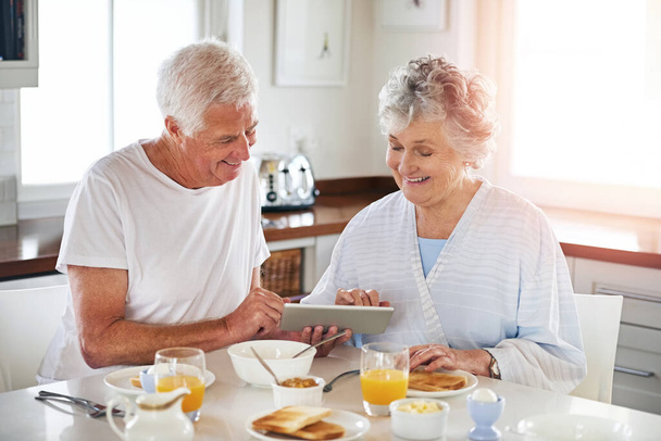 Navegar a través de múltiples sitios de noticias en lugar de un solo periódico. una pareja mayor usando una tableta digital mientras desayuna en casa - Foto, imagen