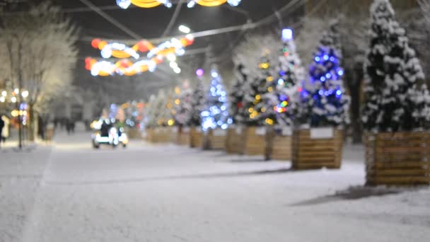 Wazige achtergrond. Mooi ingericht met verlichting en lichtgevende bloemenslingers, grote motorfiets met aanhanger rijden mensen rond versierde kerstbomen op het dorpsplein tijdens de sneeuwval op de winternacht - Video