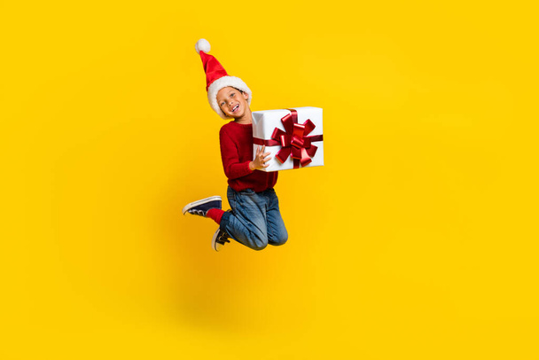 Pleine longueur photo de adorable petit garçon saut tenir cadeau énergique santa helper porter des vêtements rouges à la mode isolé sur fond de couleur jaune. - Photo, image