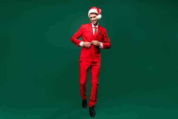 Photo of cute sweet santa claus guy dressed red jacket headwear fixing button walking isolated green color background. - 写真・画像