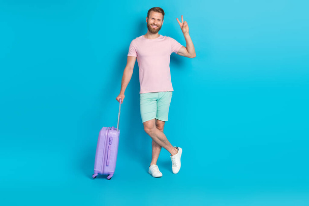 Full size photo of attractive young man travel suitcase showing v-sign cheerful wear trendy pink garment isolated on cyan color background. - 写真・画像