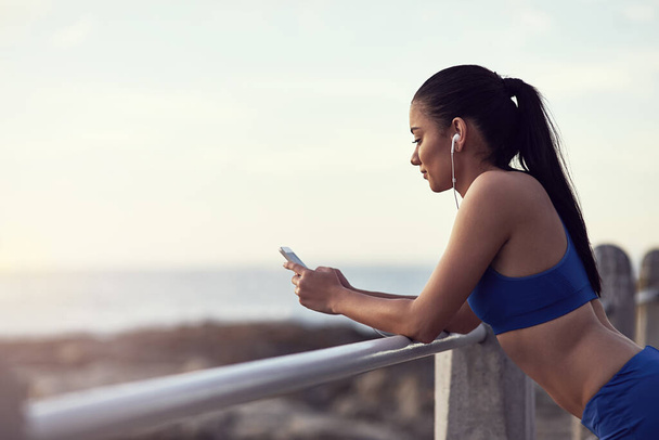 Monitoring her progress with a cool fitness app. a fit young woman using her phone and earphones during a workout outdoors - 写真・画像