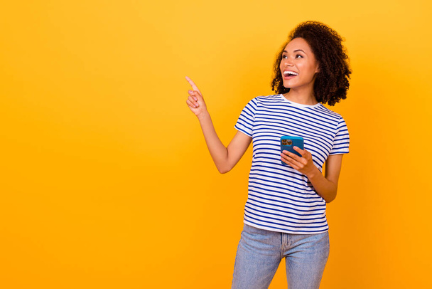 Photo of adorable excited lady wear white t-shirt looking pointing empty space typing modern device isolated yellow color background. - Foto, afbeelding