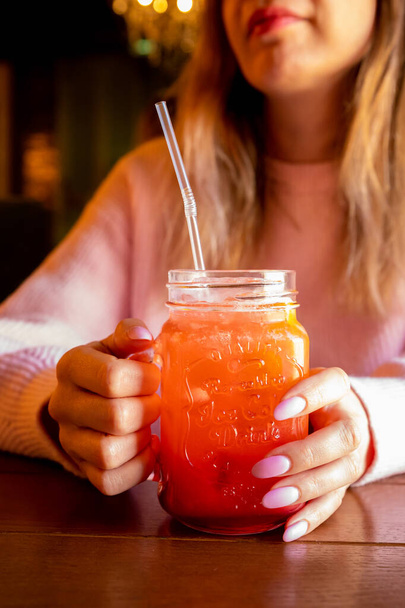 Las manos de la mujer joven caucásica sostienen el vidrio con mango de limonada de fresa roja con hielo y bebiendo paja en la mesa de madera. Manos femeninas con manicura rosa y cóctel. Vista frontal, primer plano, estilo de vida - Foto, Imagen