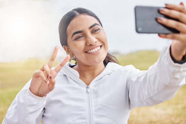Woman runner, phone selfie and peace sign in nature, park or grass for social media after exercise. Girl, smartphone and smile in photo outdoor at training, running or workout in morning for web blog. - Photo, Image