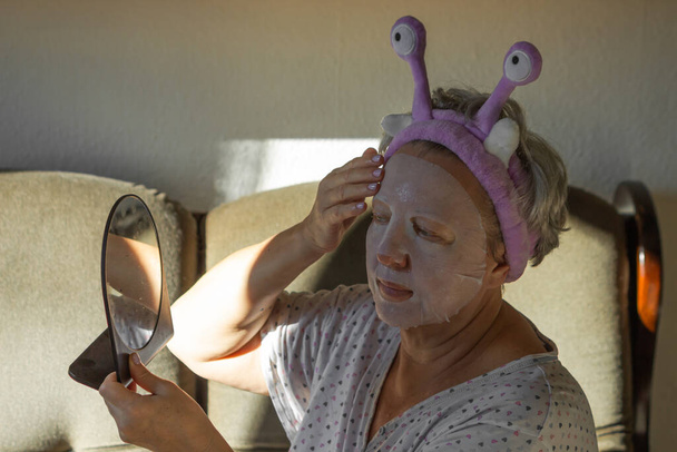 Portrait of elderly woman with funny purple headband putting on a cotton face mask at home. Lady using cosmetic mask for moisturizing facial skin, daily morning routine. Anti-aging treatments concept. - Photo, Image