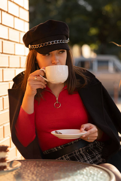 Pensive girl w stylowej czarnej czapce i jesiennym jasnoczerwonym swetrze siedzi w paryskiej kawiarni na świeżym powietrzu pijąc kawę. cieszyć się niesamowitym czasem urlopu. - Zdjęcie, obraz
