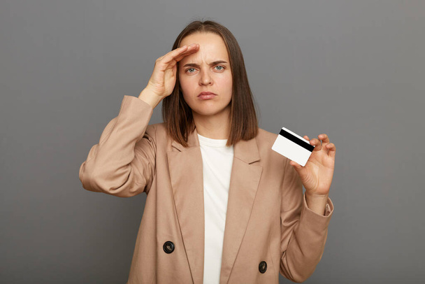 Portrait of pensive young adult brown-haired woman wearing jacket holding credit card, keeps hand near forehead, looking far, waiting her salary, standing isolated over gray background. - Foto, Imagem