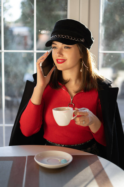 Young charming woman in red calling with cell telephone while sitting alone in coffee shop during free time, with cute smile talking conversation with mobile phone - Photo, Image