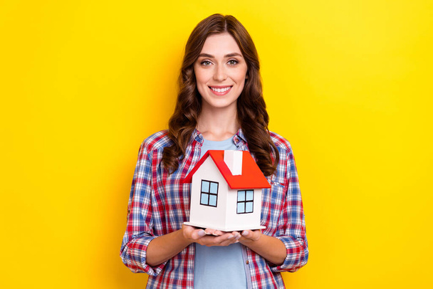Photo of adorable shiny girl dressed checkered shirt holding hands arms small red roof house isolated yellow color background. - 写真・画像