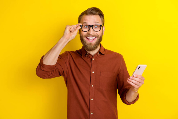 Portrait of impressed ecstatic man with masculine beard burgundy shirt hold smartphone touch eyewear isolated on yellow color background. - Fotografie, Obrázek