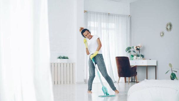 Happy housewife is doing housework and listening to music wearing modern wireless headphones, pretty young lady is singing in mop and dancing having fun at home. - 写真・画像
