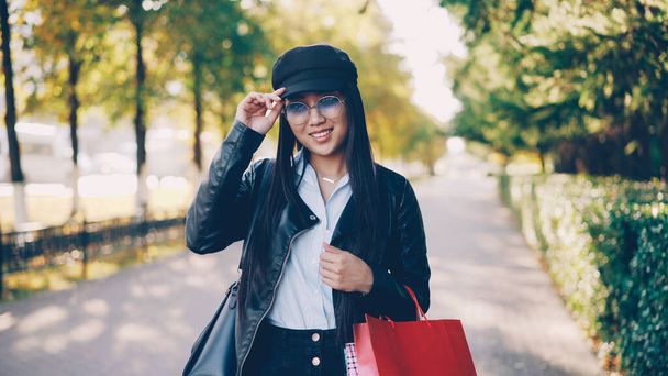 Retrato de linda chica asiática en chaqueta de cuero de moda y gorra mirando a la cámara, sonriendo y sosteniendo bolsas de papel de compras de pie al aire libre en la calle en la gran ciudad
. - Foto, Imagen