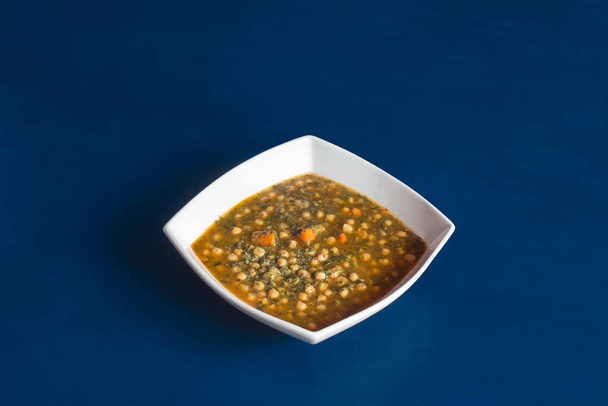 Vařená vegetariánská mísa z cizrny se špenátovou petrželkou a mrkví podávaná na bílém keramickém talíři ve tvaru hlubokého diamantu na modrém pozadí studia. - Fotografie, Obrázek