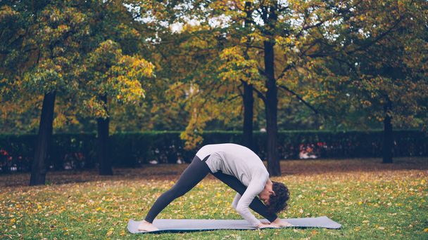 Beautiful girl with short curly hair is doing yoga outdoors stretching body and legs during individual practice in park. Trees and grass are visible. - Φωτογραφία, εικόνα