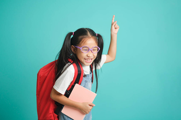 Porträt eines glücklichen kleinen Mädchens mit Rucksack über dem erhobenen Zeigefinger mit erfolgreicher Idee. Aufgeregt und glücklich. Nummer one.isolated auf blauem Pastellhintergrund. Bildungskonzept für die Schule - Foto, Bild