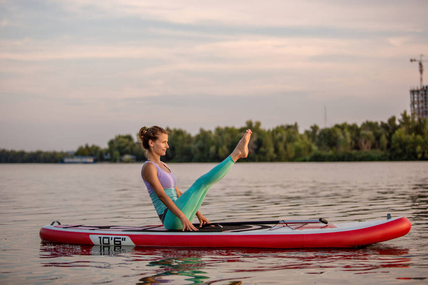 Young woman sitting on paddle board, practicing yoga pose. Doing yoga exercise on sup board, active summer rest. Exercise for flexibility and stretching of muscles. - Foto, Imagen