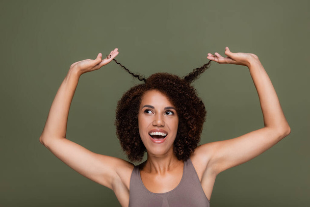 Pozitív afro-amerikai nő megérinti göndör haj és felnéz elszigetelt zöld  - Fotó, kép