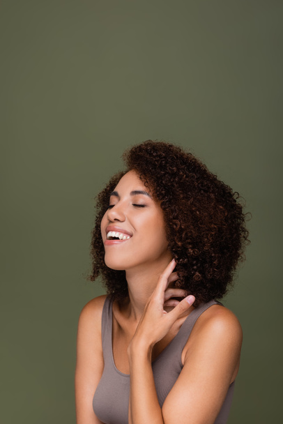 Cheerful african american woman in top touching curly hair and closing eyes isolated on green  - Foto, Imagem