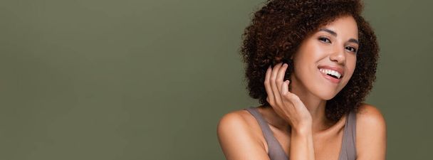 Young african american woman looking at camera while touching curly hair isolated on green, banner  - Фото, изображение
