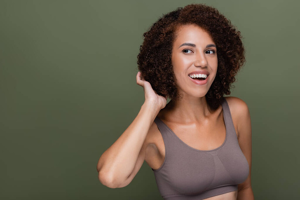 Uśmiechnięta Afroamerykanka na górze dotyka kręconych włosów i odwraca wzrok odizolowana na zielono  - Zdjęcie, obraz