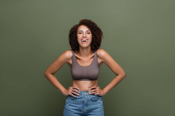 Positive african american woman in jeans and top holding hands on hips isolated on green  - Foto, Bild