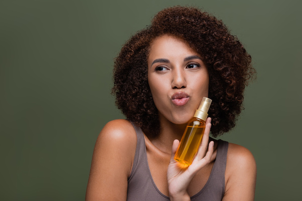 Jeune femme afro-américaine tenant de l'huile cosmétique pour les cheveux bouclés et les lèvres boudantes isolées sur vert  - Photo, image