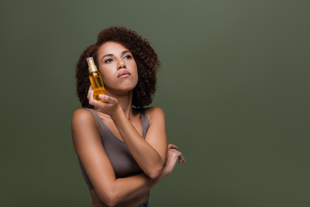 Young african american woman holding cosmetic oil and looking away isolated on green  - Foto, Imagen