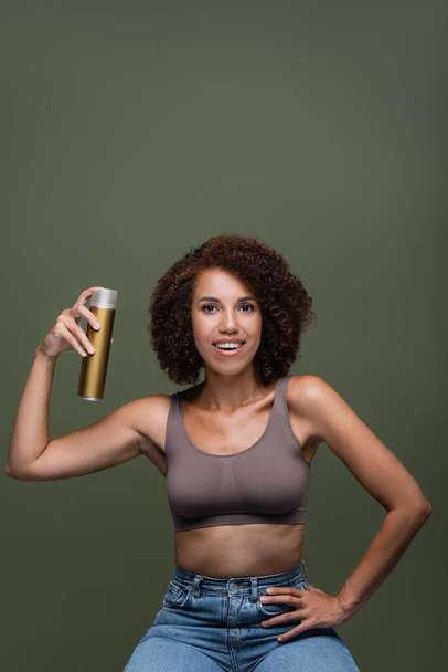 Pretty african american woman holding hairspray and looking at camera isolated on green  - Photo, Image