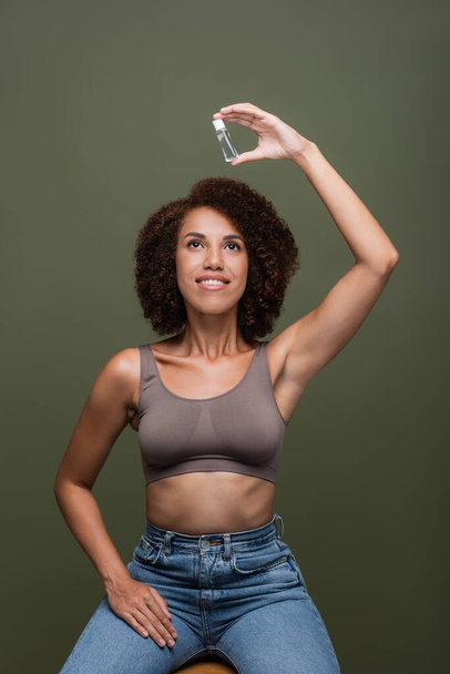 Positive african american woman in jeans and holding looking at bottle of cosmetic oil isolated on green  - Photo, image