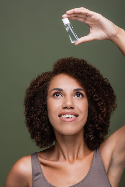 Hübsche und lockige afrikanisch-amerikanische Frau beim Betrachten von Kosmetiköl isoliert auf grün  - Foto, Bild