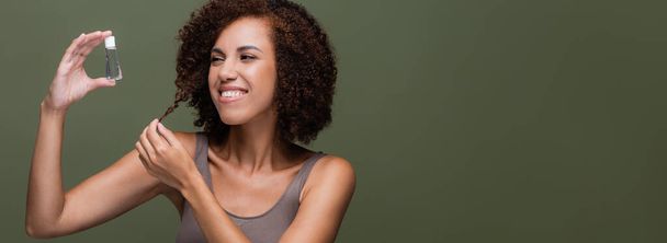 Cheerful african american woman looking at cosmetic oil and touching curly hair isolated on green, banner  - Foto, imagen