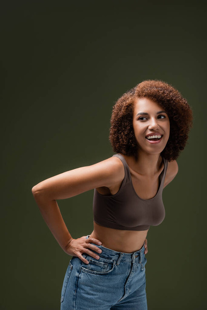 Positieve Afro-Amerikaanse vrouw in top en jeans op zoek weg geïsoleerd op donkergroen  - Foto, afbeelding