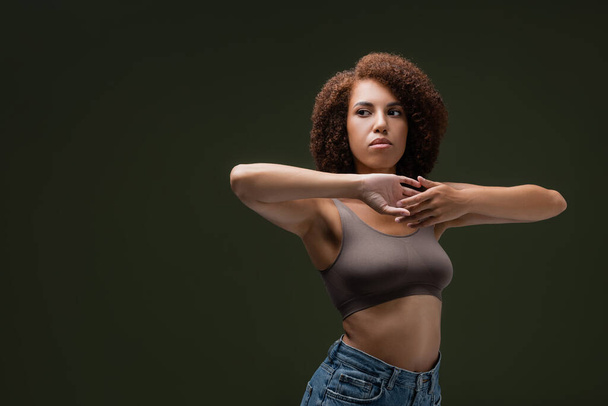 Young and curly african american model in top touching hands isolated on dark green  - Foto, Imagem